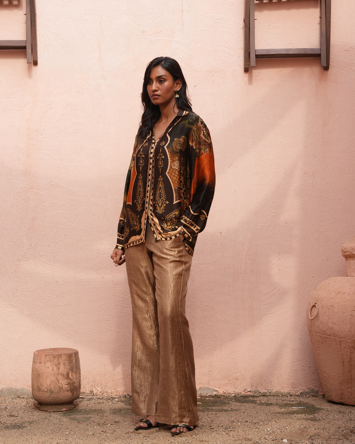 A woman stands against a textured pink wall wearing a bohemian-style patterned jacket with metallic gold wide-leg pants and sandals.