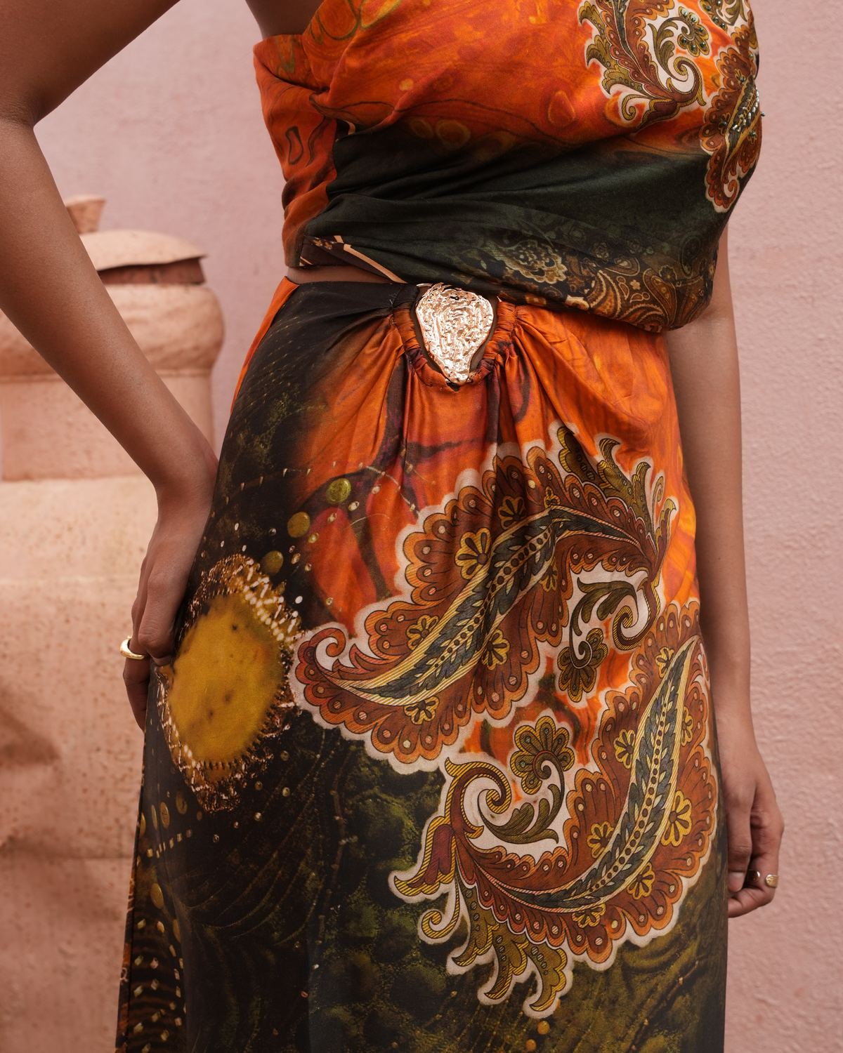 A close-up of a model wearing a draped orange and black printed fabric garment with ornate paisley and floral motifs, tied at the waist with a decorative metallic ornament.