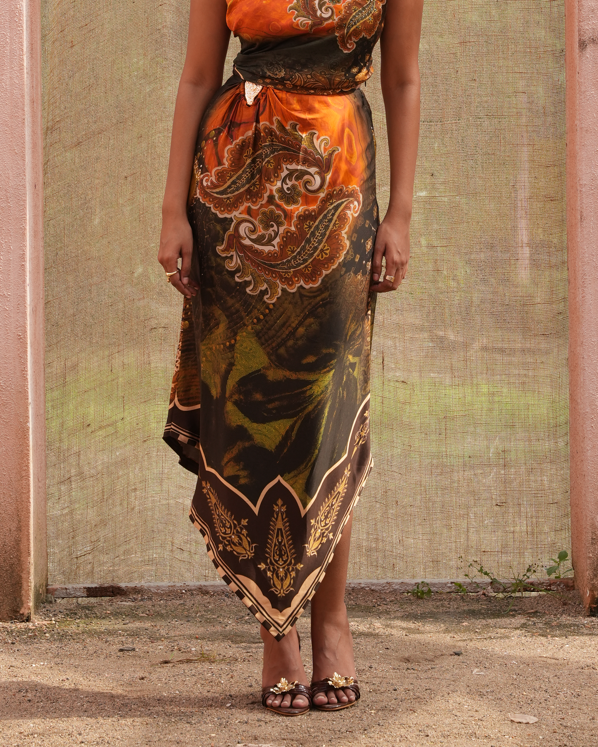 A woman wearing a sleeveless wrap-style maxi dress with a vibrant orange, brown, and green paisley print, standing outdoors against a rustic beige wall.
