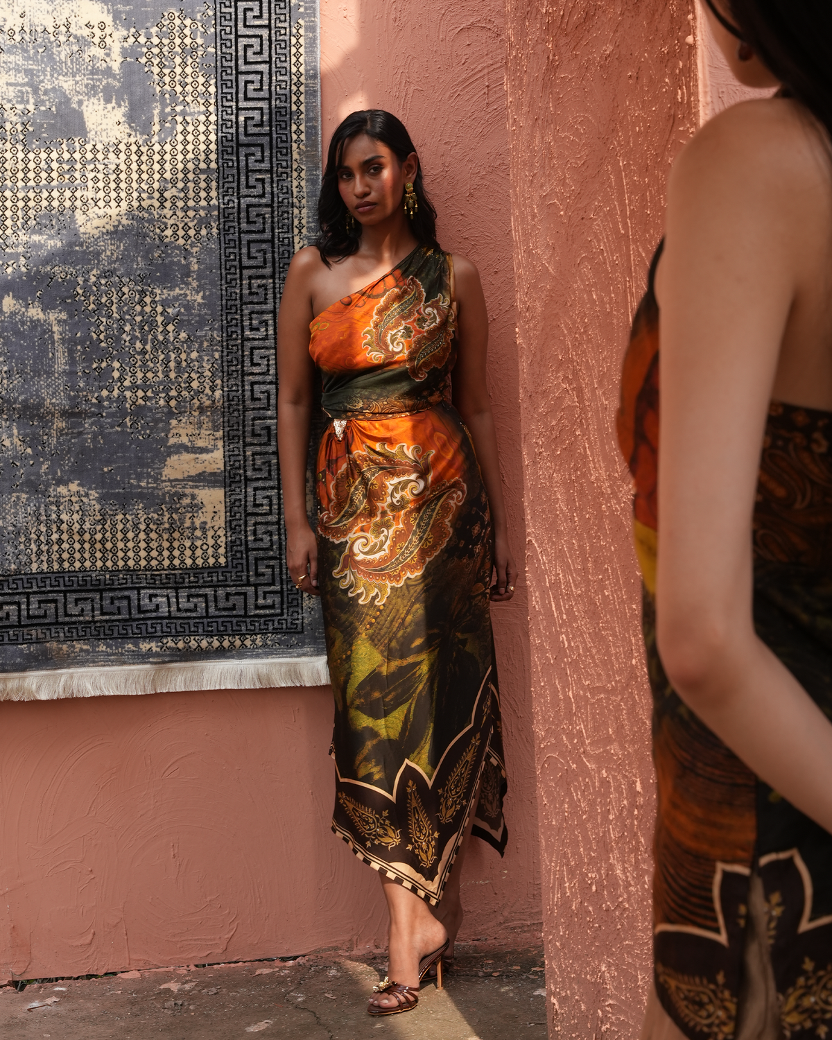 A woman models a one-shoulder silk dress with a vibrant orange, green, and gold paisley pattern, standing against a textured terracotta wall next to a patterned rug.