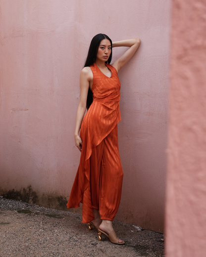 A woman wearing a sleeveless rust-orange draped maxi dress and gold heeled sandals, posing against a textured pink stucco wall.