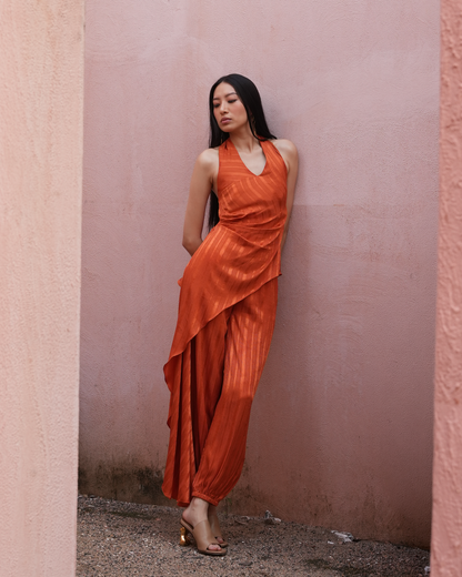A woman with long black hair stands against a dusty pink wall wearing a sleeveless, draped orange dress with pleated wide-leg pants and gold heeled sandals.