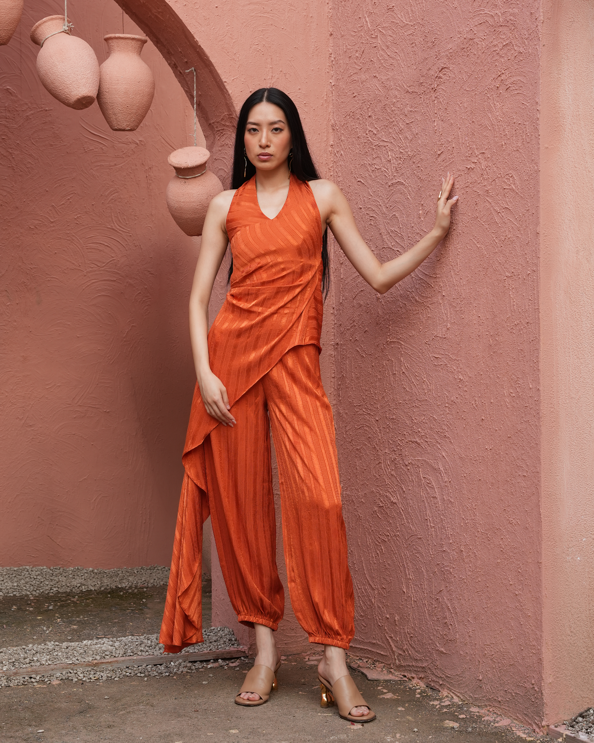A woman stands against a textured pink wall wearing a sleeveless draped orange jumpsuit with matching pleated pants and clear high-heeled sandals.