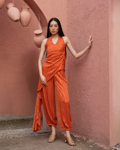A woman stands against a textured pink wall wearing a sleeveless draped orange jumpsuit with matching pleated pants and clear high-heeled sandals.