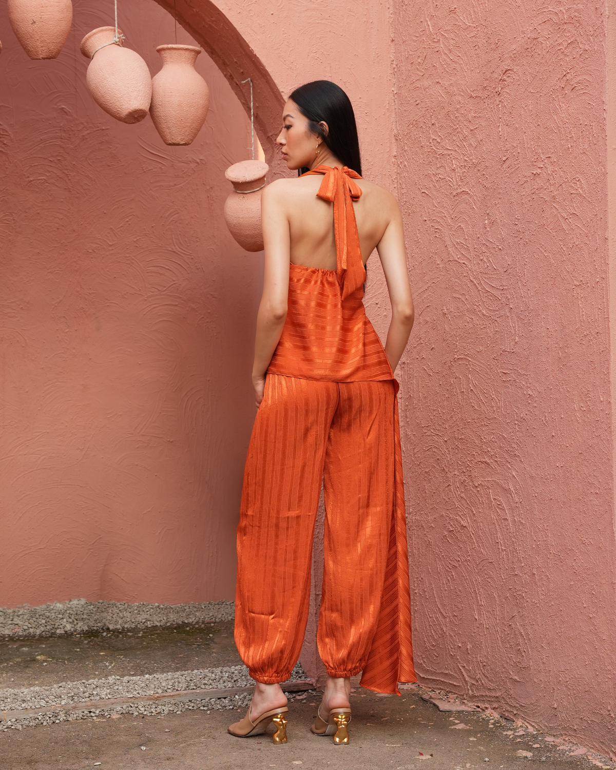 A woman stands with her back to the camera wearing a bright orange halter-neck jumpsuit with pleated wide-leg pants and gold heels, posed against a textured pink wall with hanging terracotta pots.