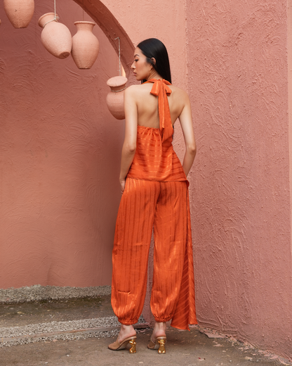 A woman stands with her back to the camera wearing a bright orange halter-neck jumpsuit with pleated wide-leg pants and gold heels, posed against a textured pink wall with hanging terracotta pots.