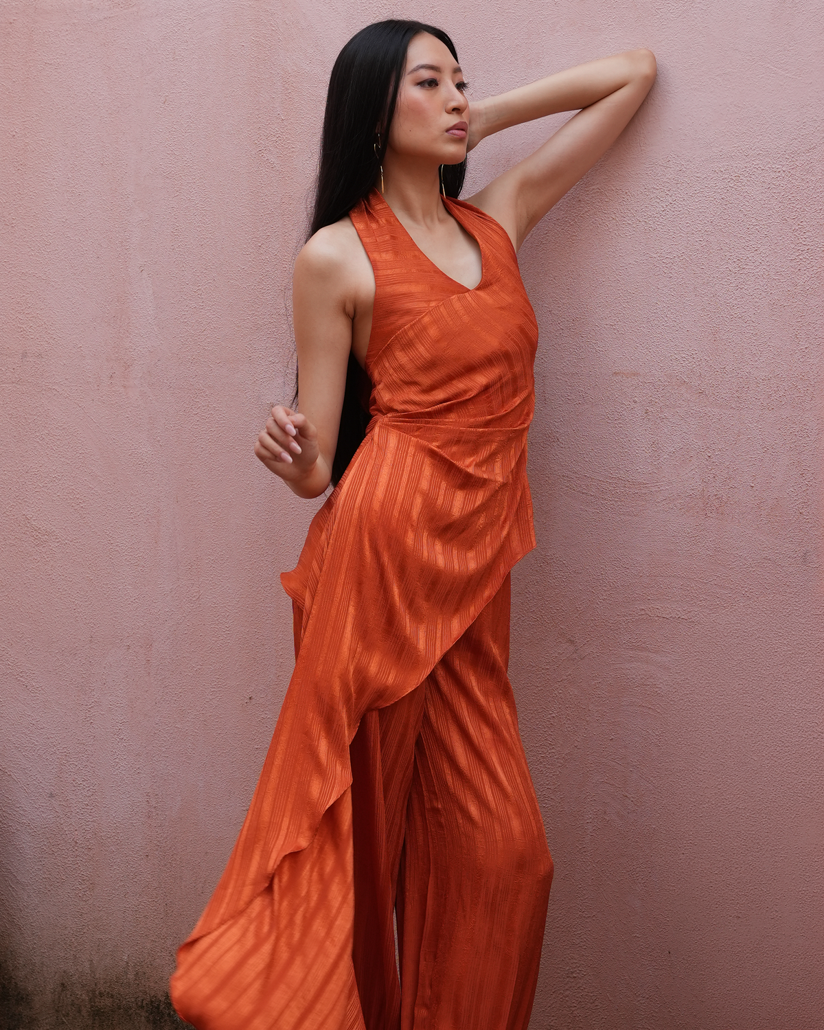 A woman wearing a sleeveless, draped orange jumpsuit with wide-leg pants, posing against a textured pink wall.
