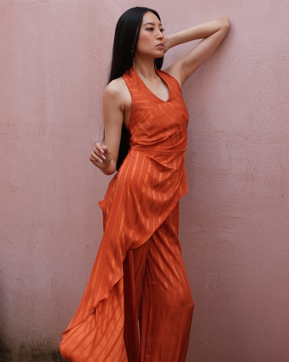 A woman wearing a sleeveless, draped orange jumpsuit with wide-leg pants, posing against a textured pink wall.