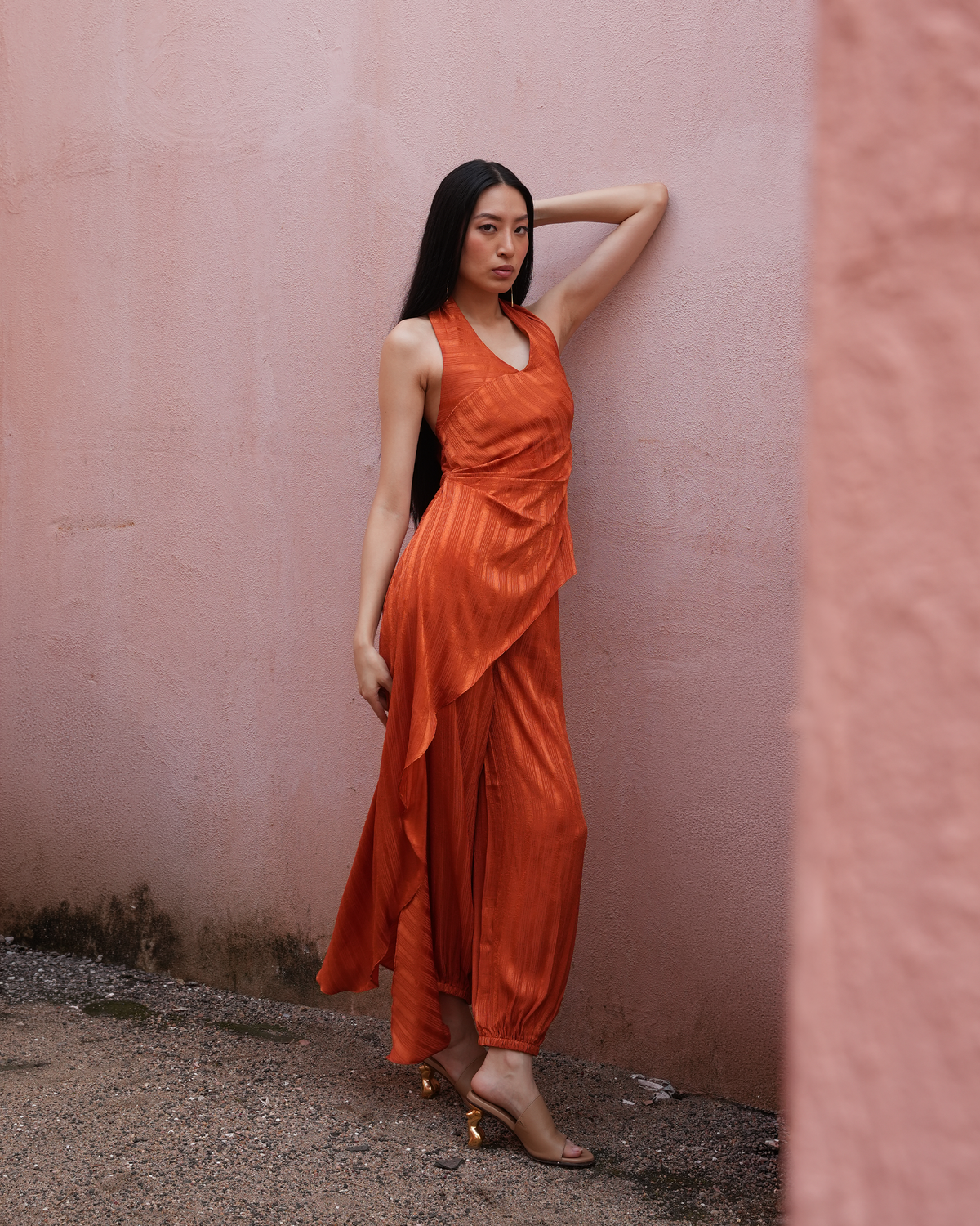A woman wearing a sleeveless rust-orange draped maxi dress and gold heeled sandals, posing against a textured pink stucco wall.