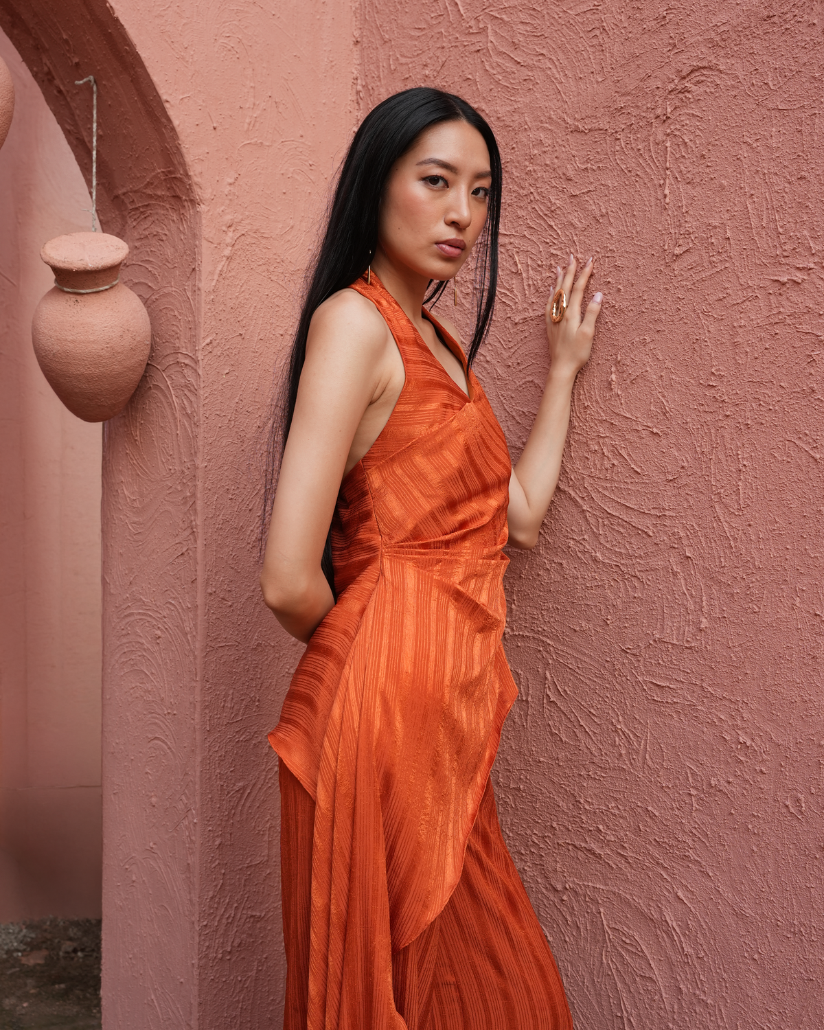 A woman with long black hair stands against a textured terracotta-pink stucco wall wearing a sleeveless draped orange dress. She is posing with one hand on the wall, with a hanging clay pot visible on the left.