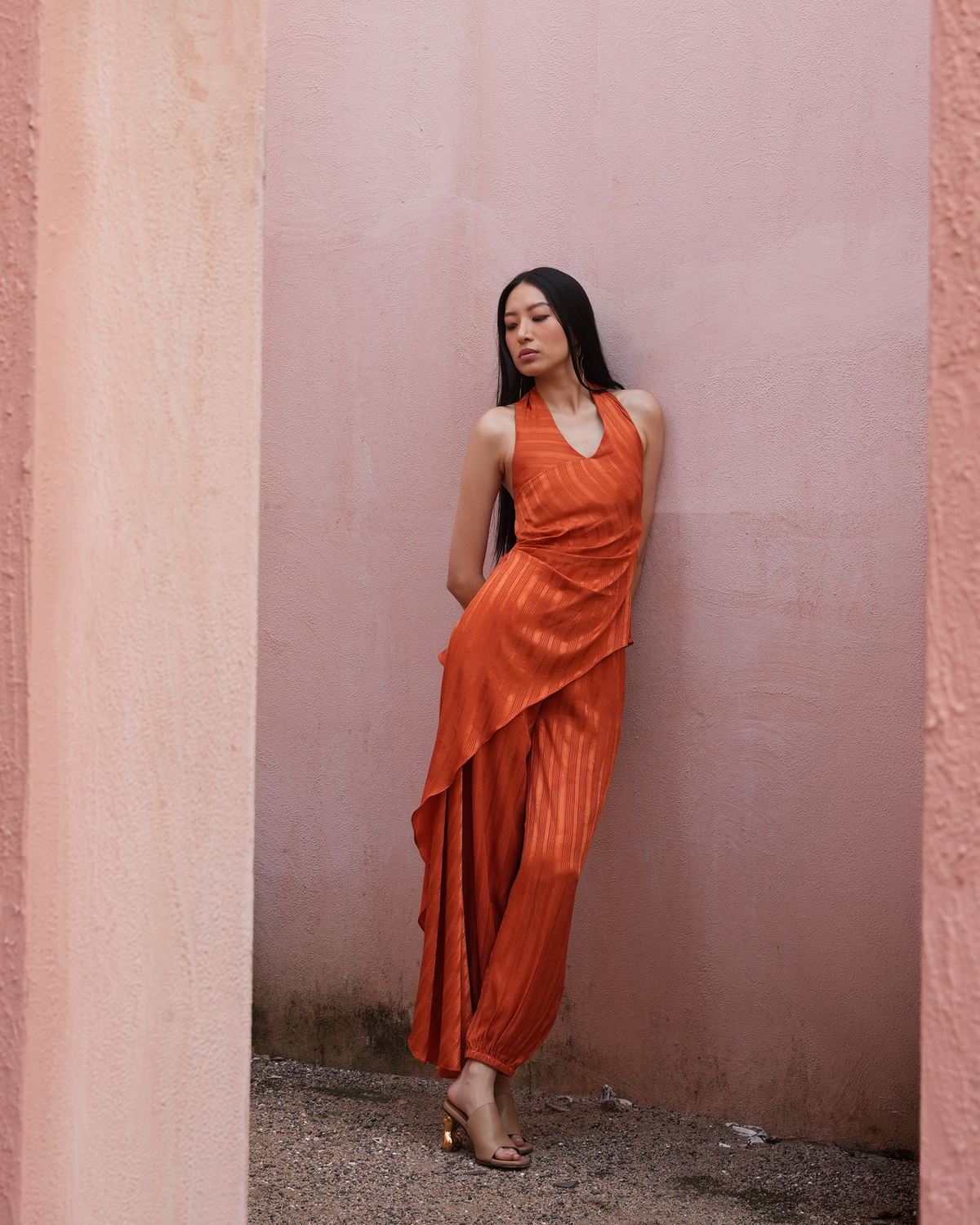 A woman wearing a shimmering orange halter dress with draped layers and gold heeled sandals, posing against a textured pink wall.