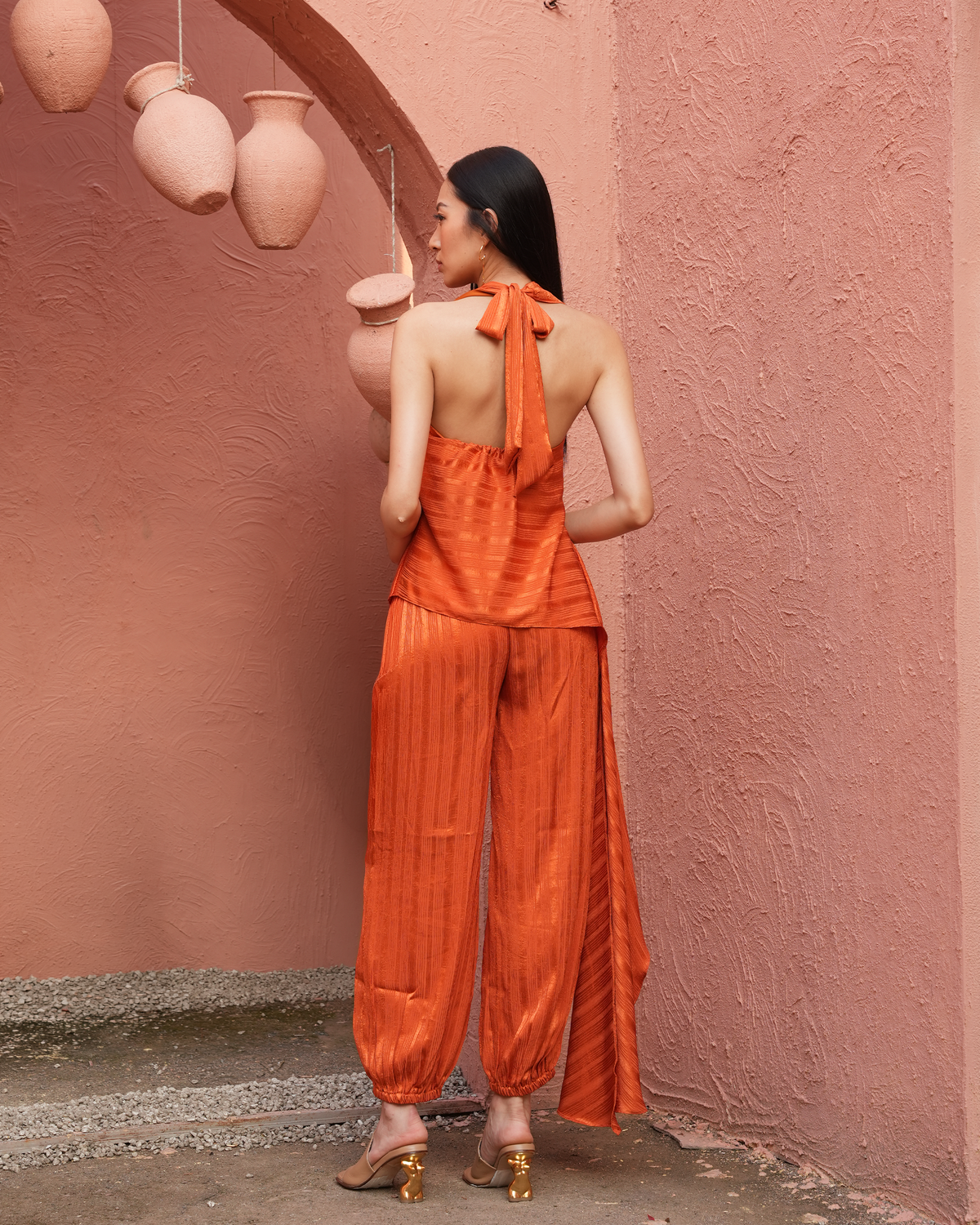 A woman stands with her back to the camera wearing a bright orange satin halter top and matching pleated wide-leg pants, posed against a textured terracotta wall with hanging clay pots.