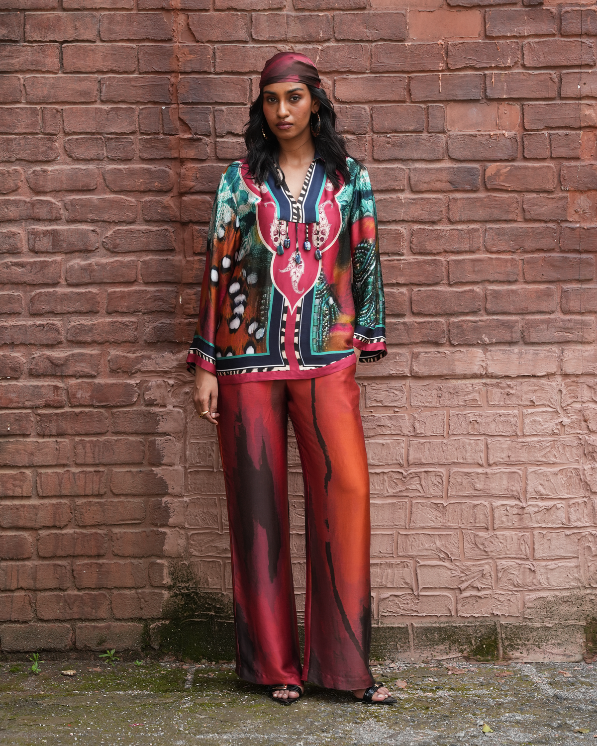 A woman stands against a red-brown brick wall wearing a matching printed outfit with a long-sleeve top, wide-leg pants, and a headscarf. The outfit features a bold multicolor pattern with red, orange, teal, black, and white elements.