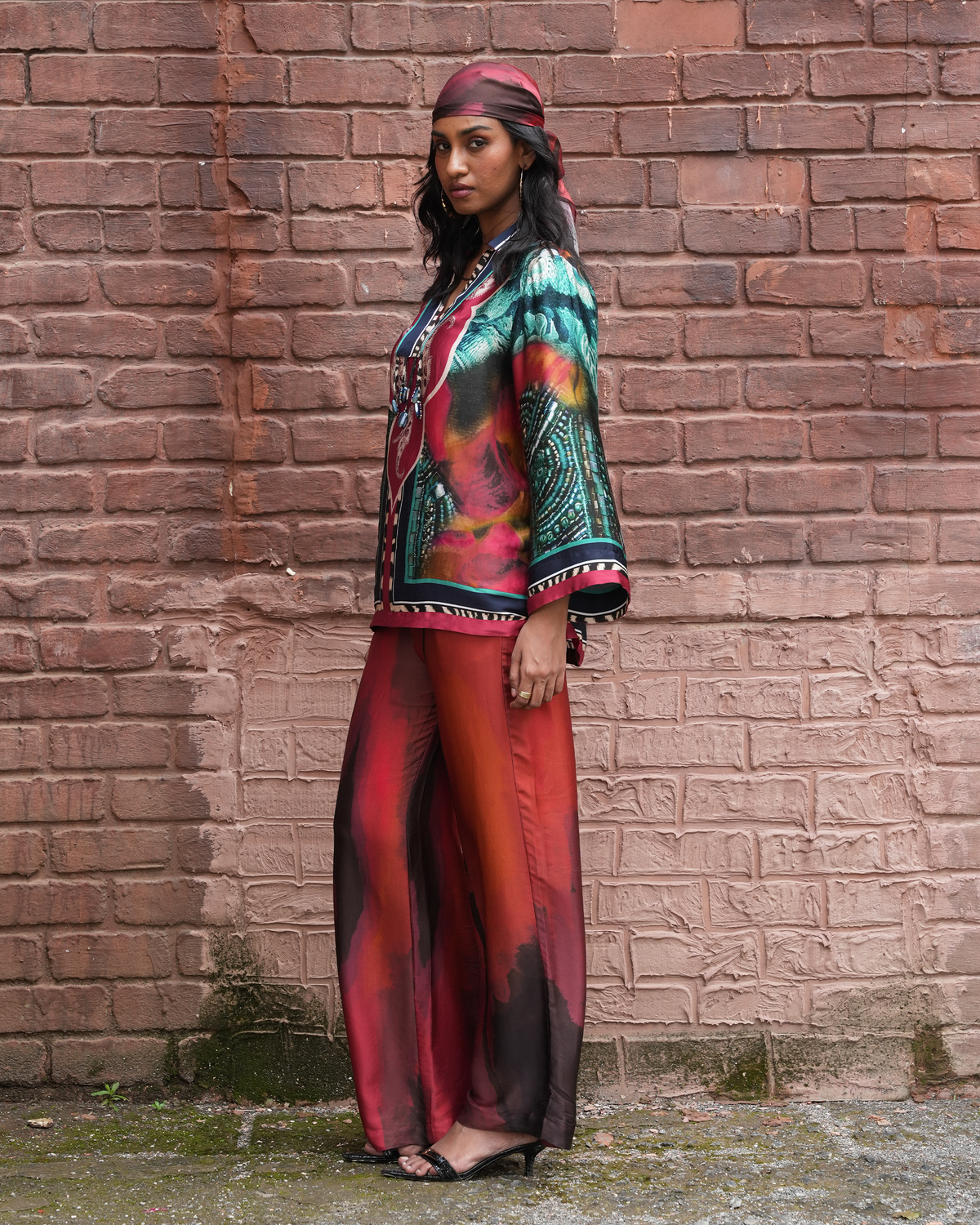 A woman stands in profile against a weathered red-brown brick wall, wearing a matching printed outfit with a headscarf. The set features a vibrant multicolor print with red, teal, black, and orange tones.