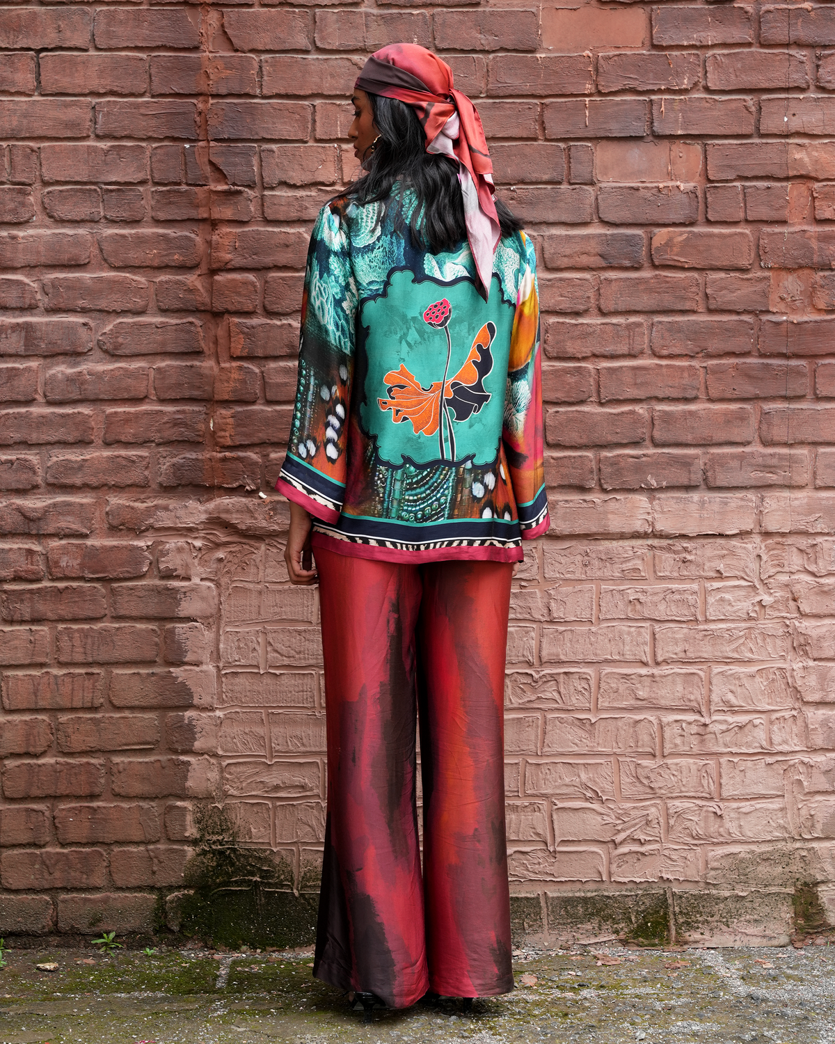 A model stands with their back to the camera in front of a rustic red brick wall, wearing a vibrant multicolor printed jacket with a butterfly design, matching red wide-leg pants, and a pink headscarf.