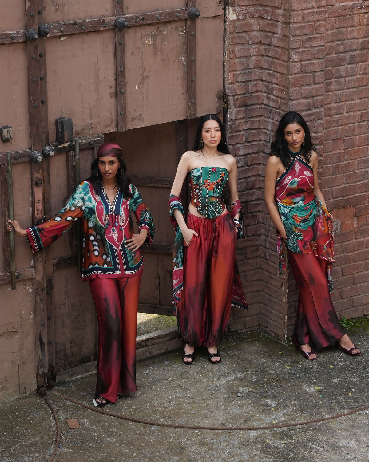 Three women modeling matching bohemian-style printed outfits in front of a rustic brick and wood wall. The garments feature bold multicolor patterns with red, teal, green, black, and white, including a long-sleeve top and wide-leg pants on two models, and a strapless corset-style top with matching draped pants on the center model.