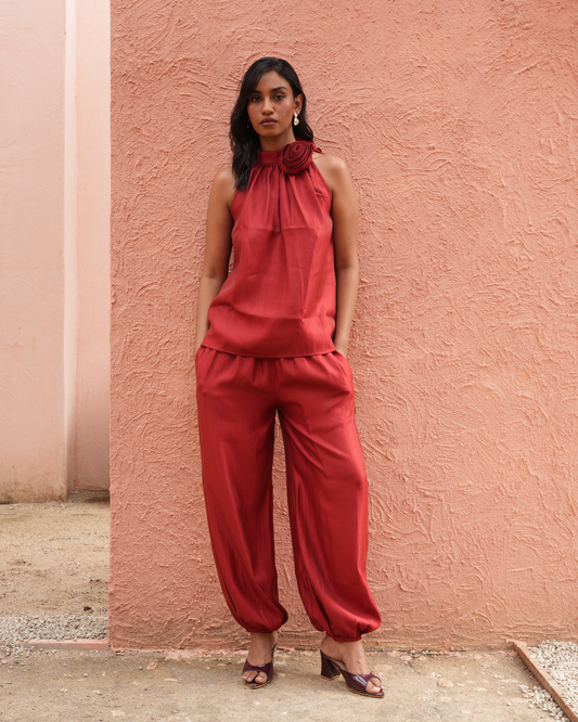 A woman stands against a textured terracotta wall wearing a sleeveless rust-red halter top with a large gathered neckline and matching loose harem-style pants, paired with dark brown heeled sandals.