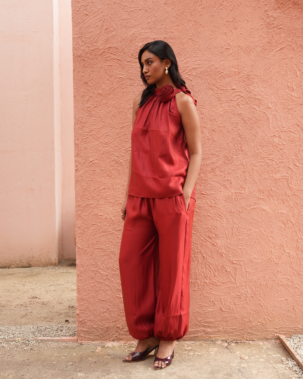 A woman stands against a textured peach-pink stucco wall wearing a matching red sleeveless halter top and wide-leg pants set, paired with dark heeled sandals.