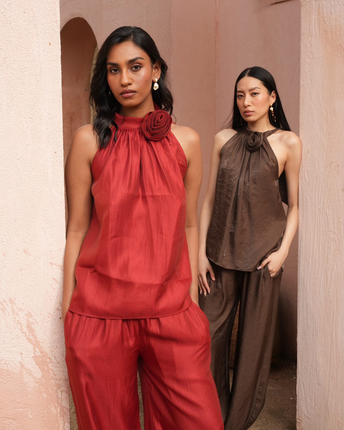Two women modeling matching draped halter-style tops and wide-leg pants in warm earth tones, standing in a textured pink architectural setting. The front model wears a rust red set, and the model behind wears a taupe-brown set.