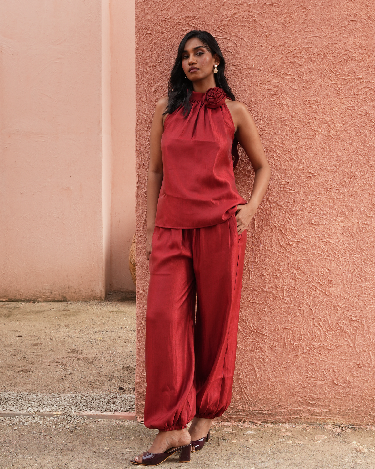 A woman stands against a textured terracotta wall wearing a matching red sleeveless top with a large fabric flower at the neckline and red wide-leg pants. She is wearing dark brown heeled sandals.