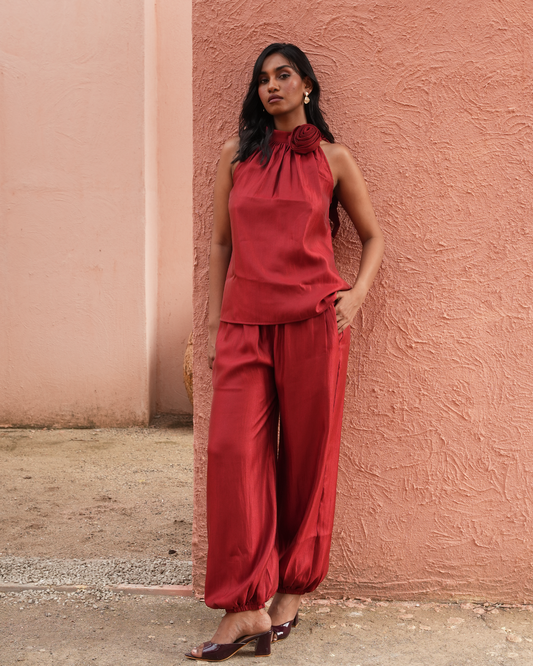 A woman stands against a textured terracotta wall wearing a matching red sleeveless top with a large fabric flower at the neckline and red wide-leg pants. She is wearing dark brown heeled sandals.