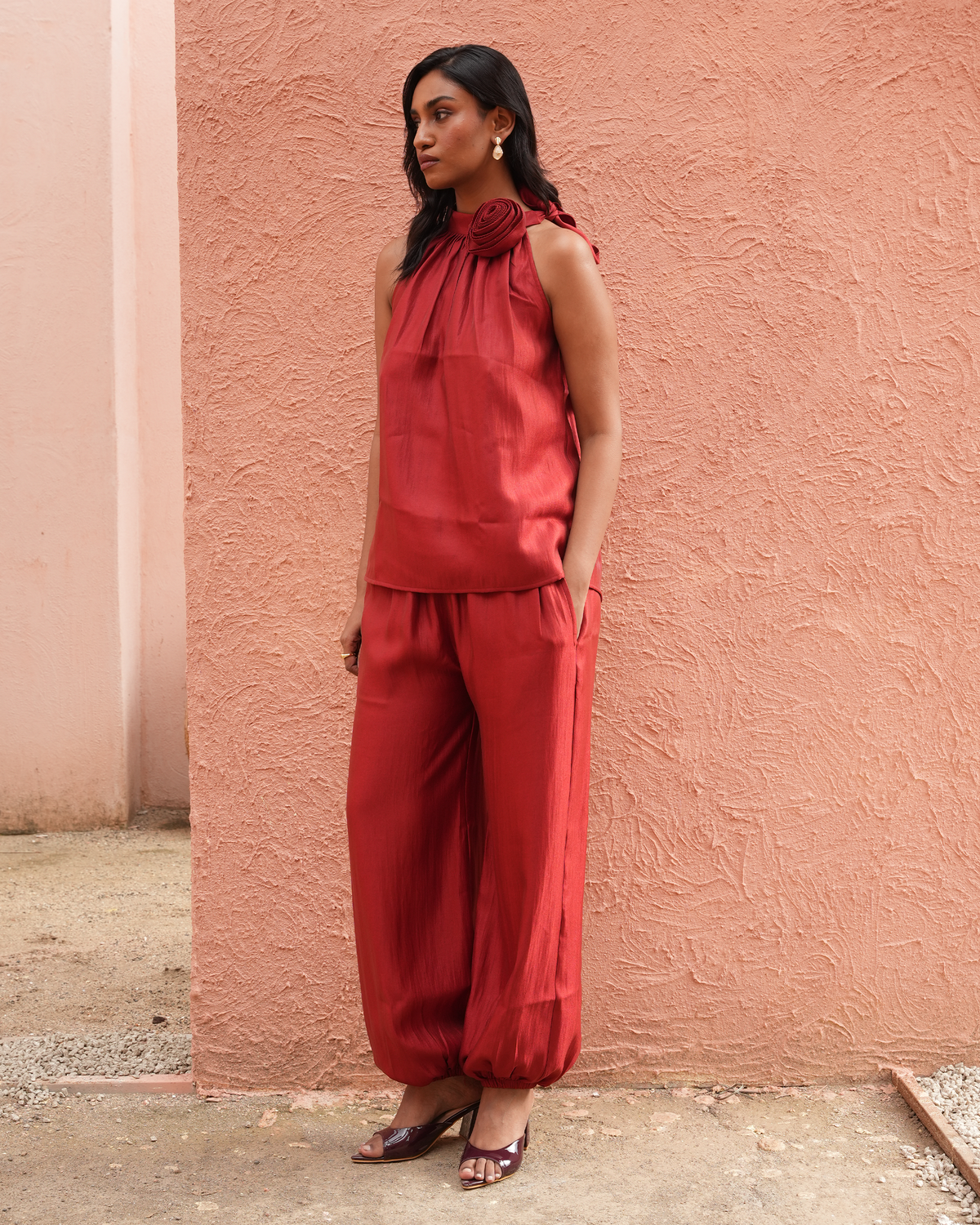 A woman stands against a textured peach-pink stucco wall wearing a matching red sleeveless top and wide-leg pants set, paired with dark heeled sandals.