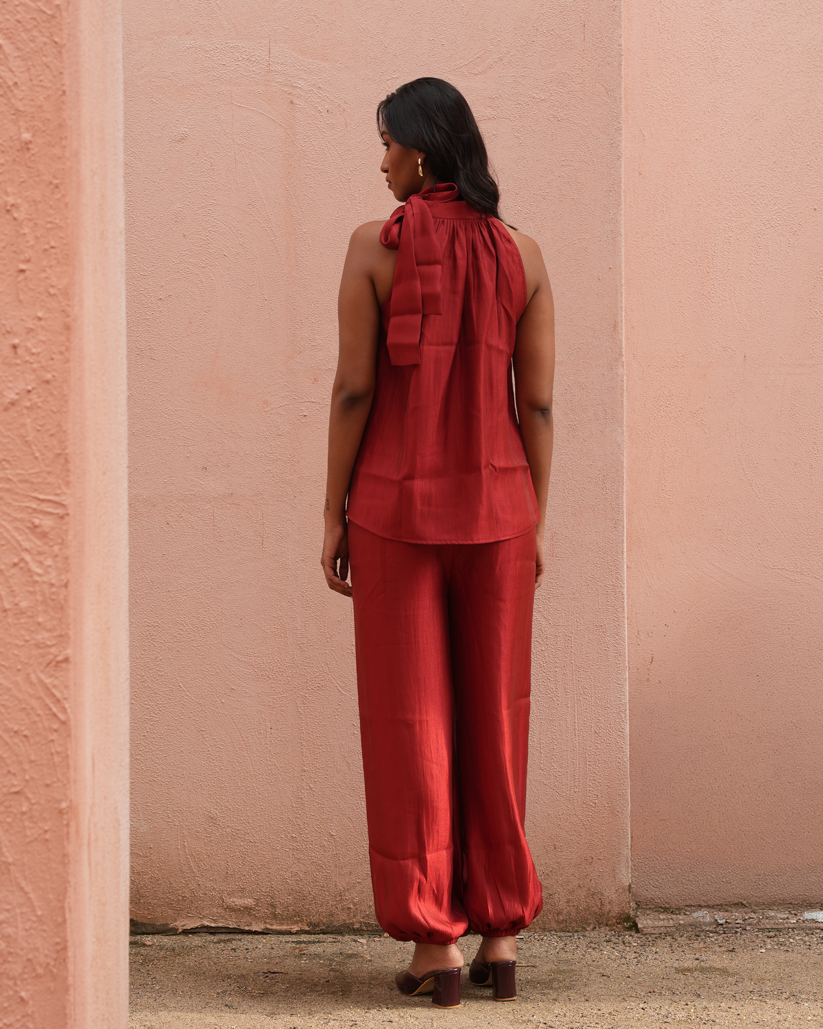 A woman stands with her back to the camera wearing a matching rust-red sleeveless top and wide-leg pants set, posed against a textured peach-pink stucco wall.