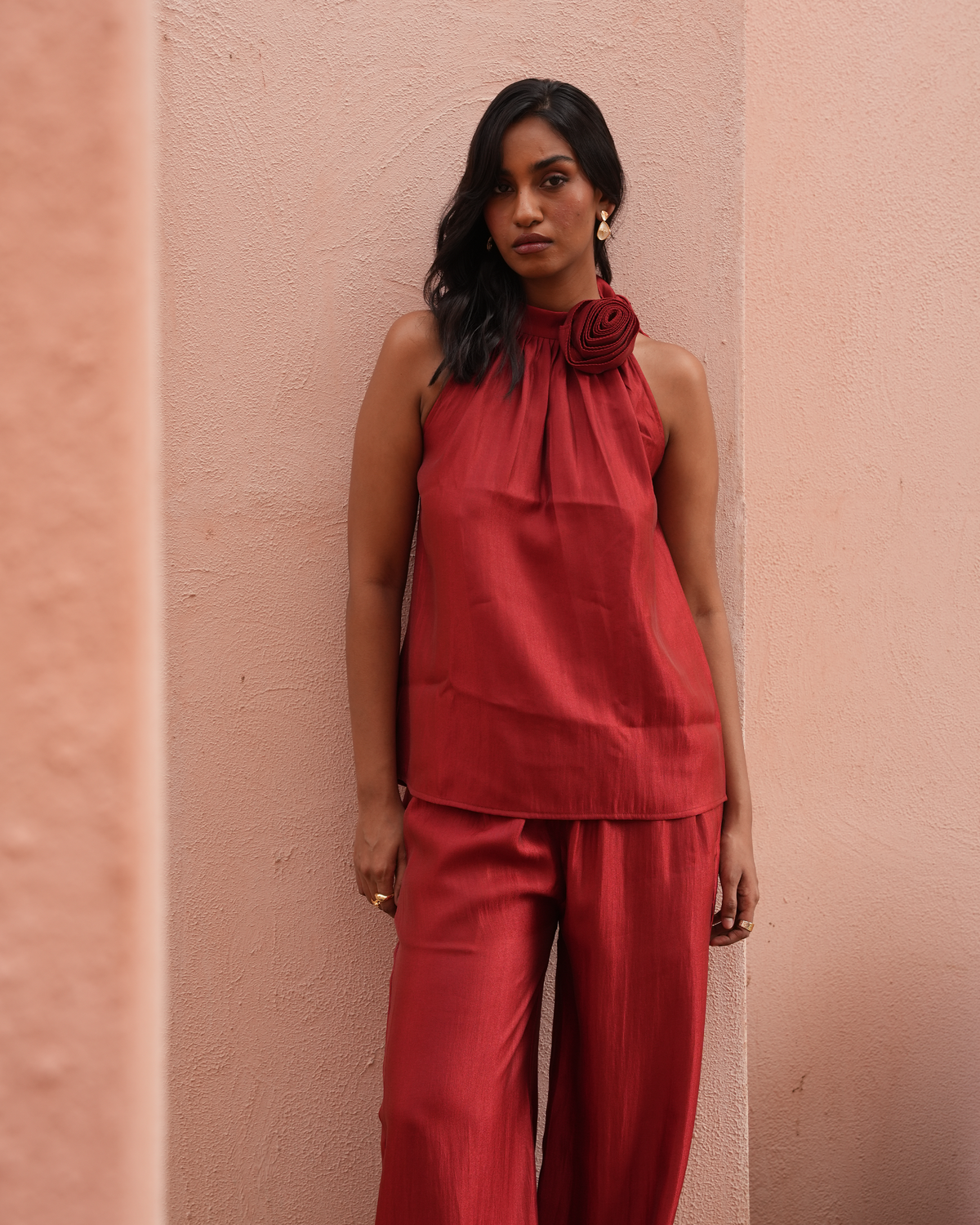 A woman wearing a matching rust-red sleeveless top with a large fabric rose detail at the collar and matching wide-leg pants, standing against a textured peach-pink wall.