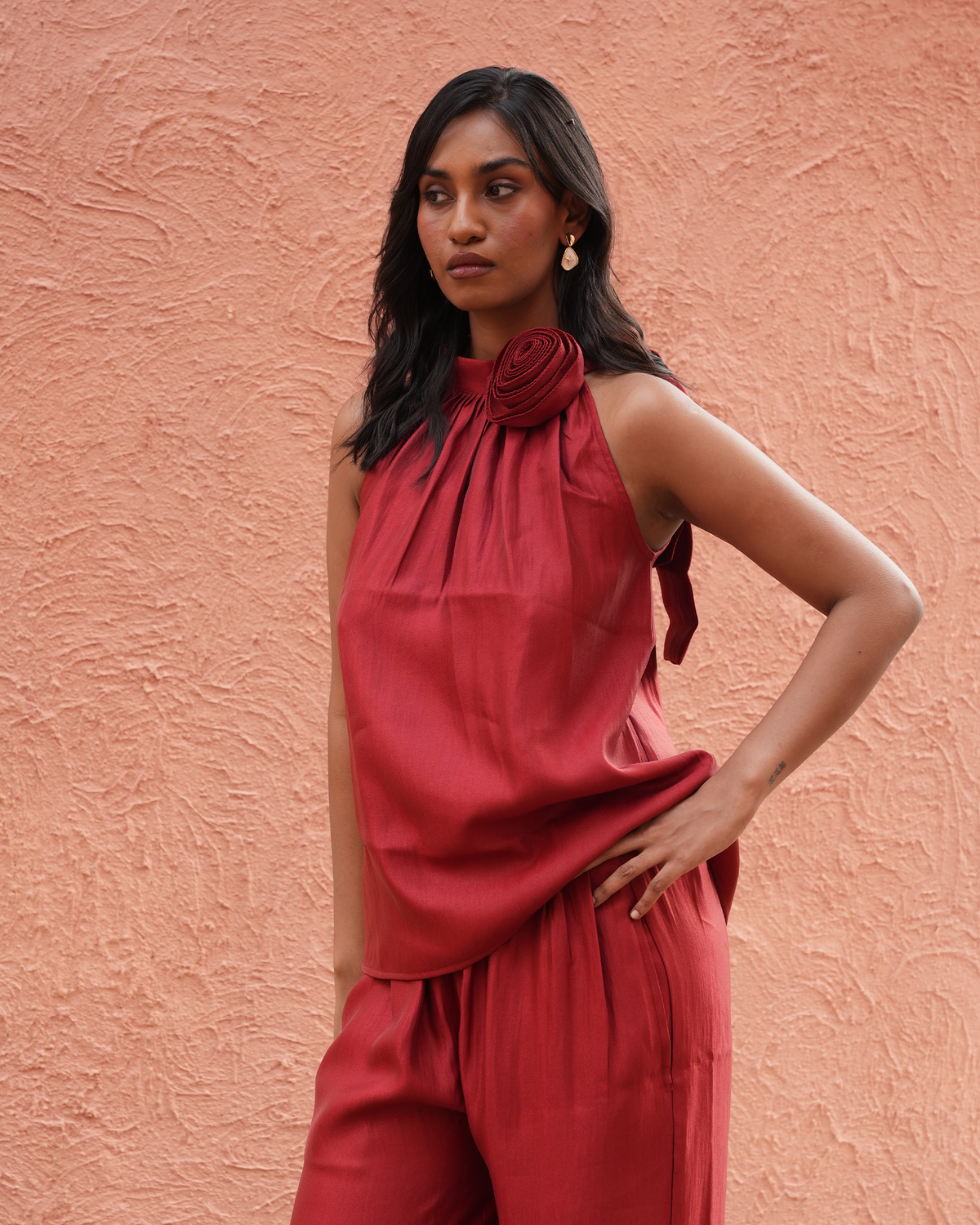 A woman wearing a matching rust-red sleeveless top and trousers set, standing against a textured terracotta wall.