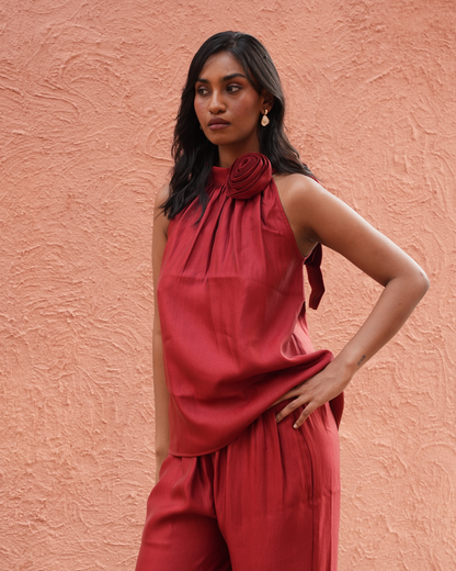 A woman wearing a matching rust-red sleeveless top and trousers set, standing against a textured terracotta wall.