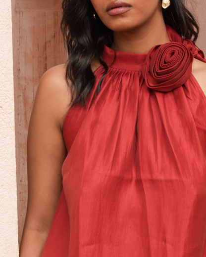 A close-up of a woman wearing a sleeveless rust-red top with a gathered high neckline and a large fabric rosette detail on the left shoulder, set against a neutral textured wall.