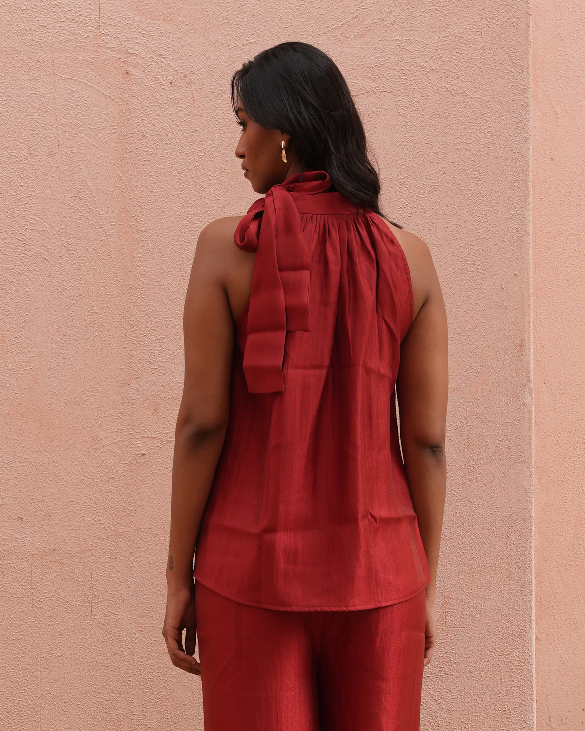 A woman is shown from the back wearing a sleeveless rust-red top with a large bow detail at the neck and matching rust-red pants, standing against a textured pink wall.