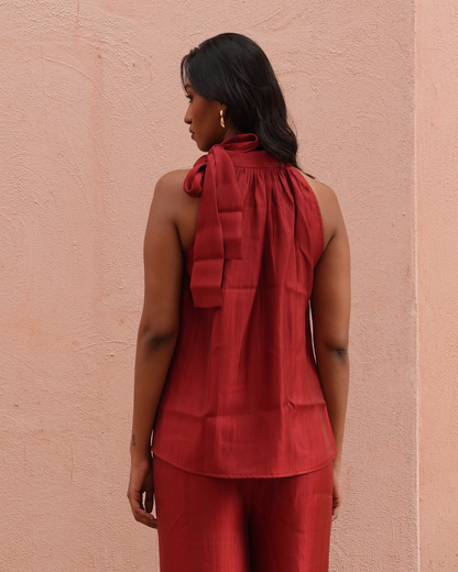 A woman is shown from the back wearing a sleeveless rust-red top with a large bow detail at the neck and matching rust-red pants, standing against a textured pink wall.
