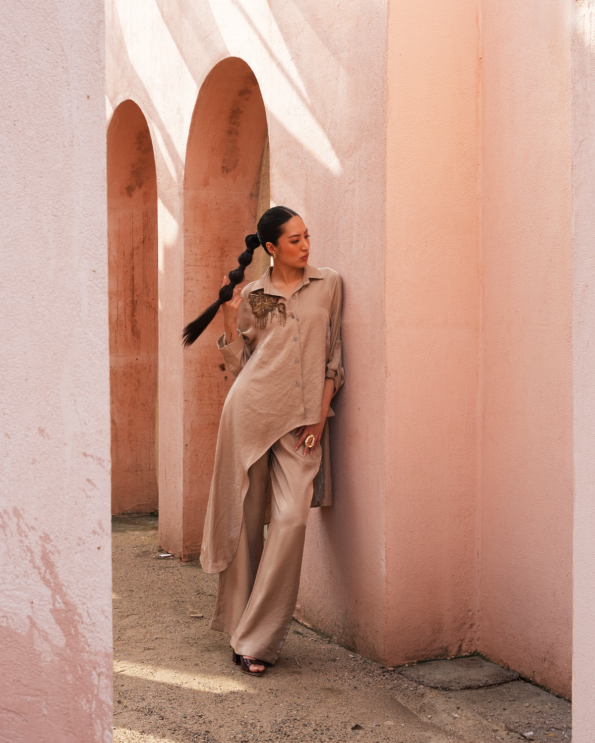 A woman with a long black braid stands in a dusty pink architectural corridor with arches, wearing a beige oversized button-up shirt, matching wide-leg trousers, and black sandals.