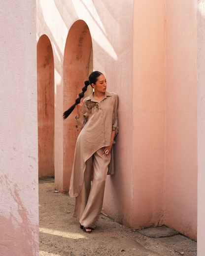 A woman with a long black braid stands in a dusty pink architectural corridor with arches, wearing a beige oversized button-up shirt, matching wide-leg trousers, and black sandals.