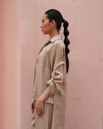 A woman in profile wearing a light beige linen shirt and matching trousers, standing against a textured pink wall.