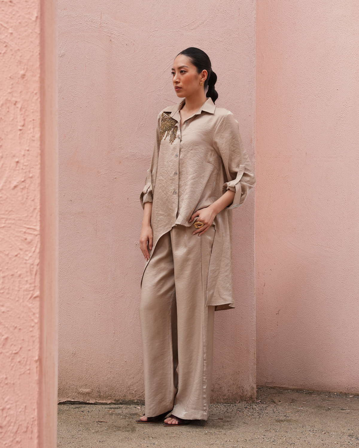 A woman stands in a pink textured outdoor setting wearing a matching beige linen shirt and wide-leg trousers set.