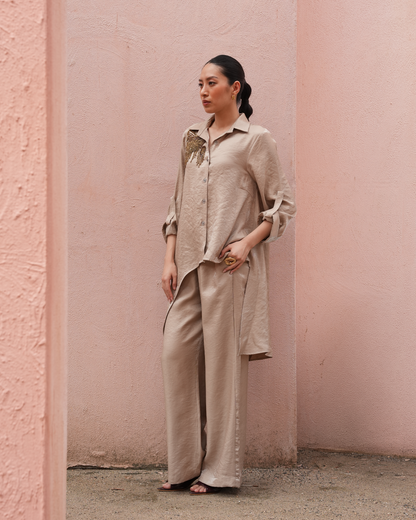 A woman stands in a pink textured outdoor setting wearing a matching beige linen shirt and wide-leg trousers set.