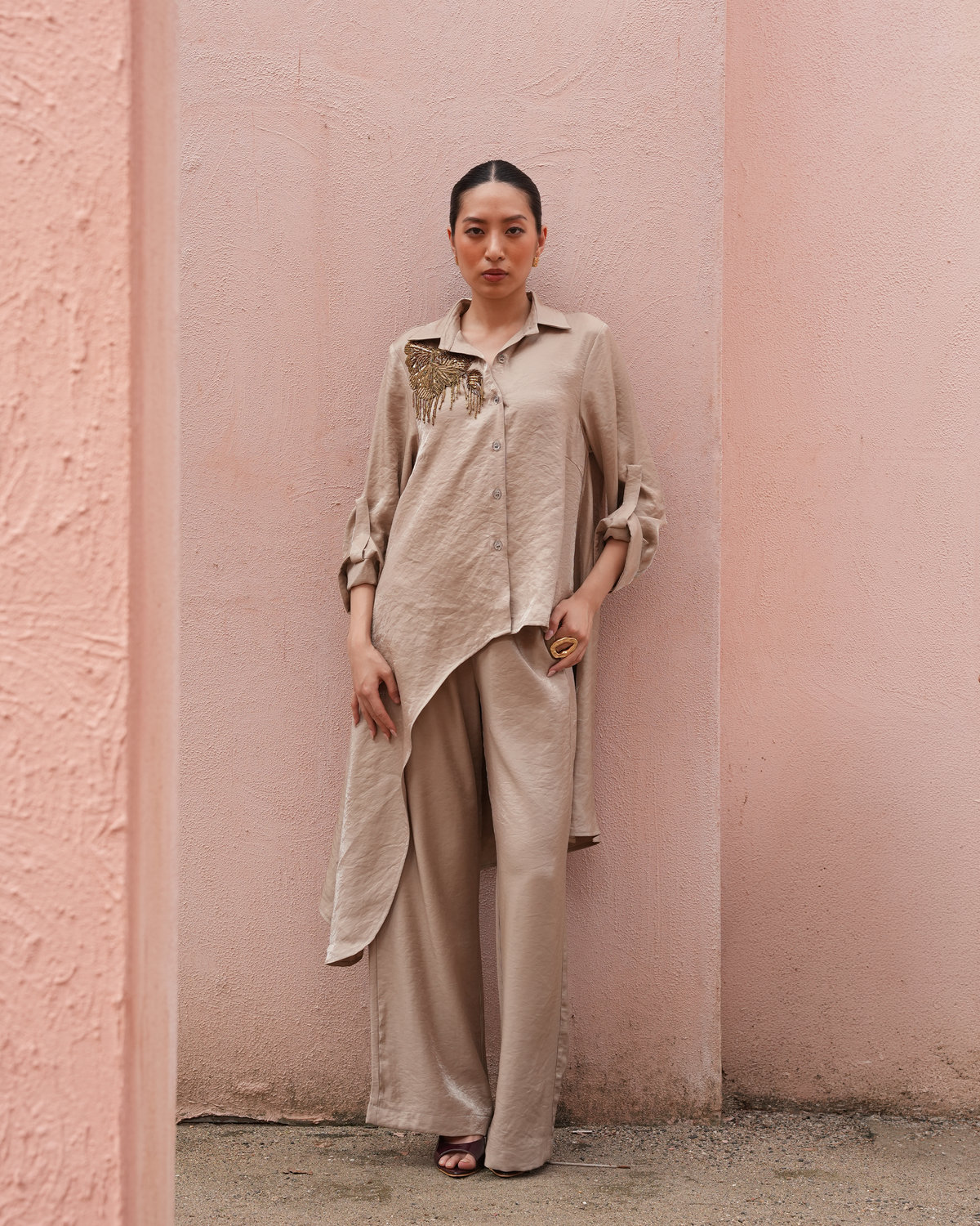 A woman stands against a pink textured wall wearing a loose, asymmetrical beige linen shirt with gold embroidery on the chest, matching beige wide-leg pants, and dark sandals.