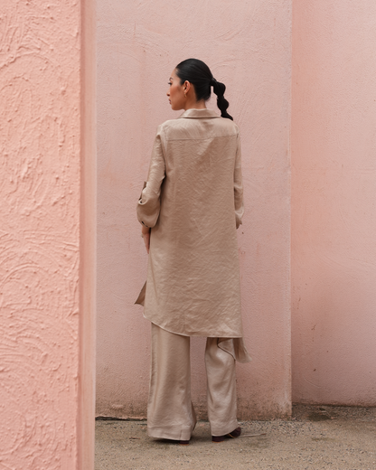 A woman stands with her back to the camera wearing a light beige linen long coat and matching wide-leg trousers, set against a textured pink wall.