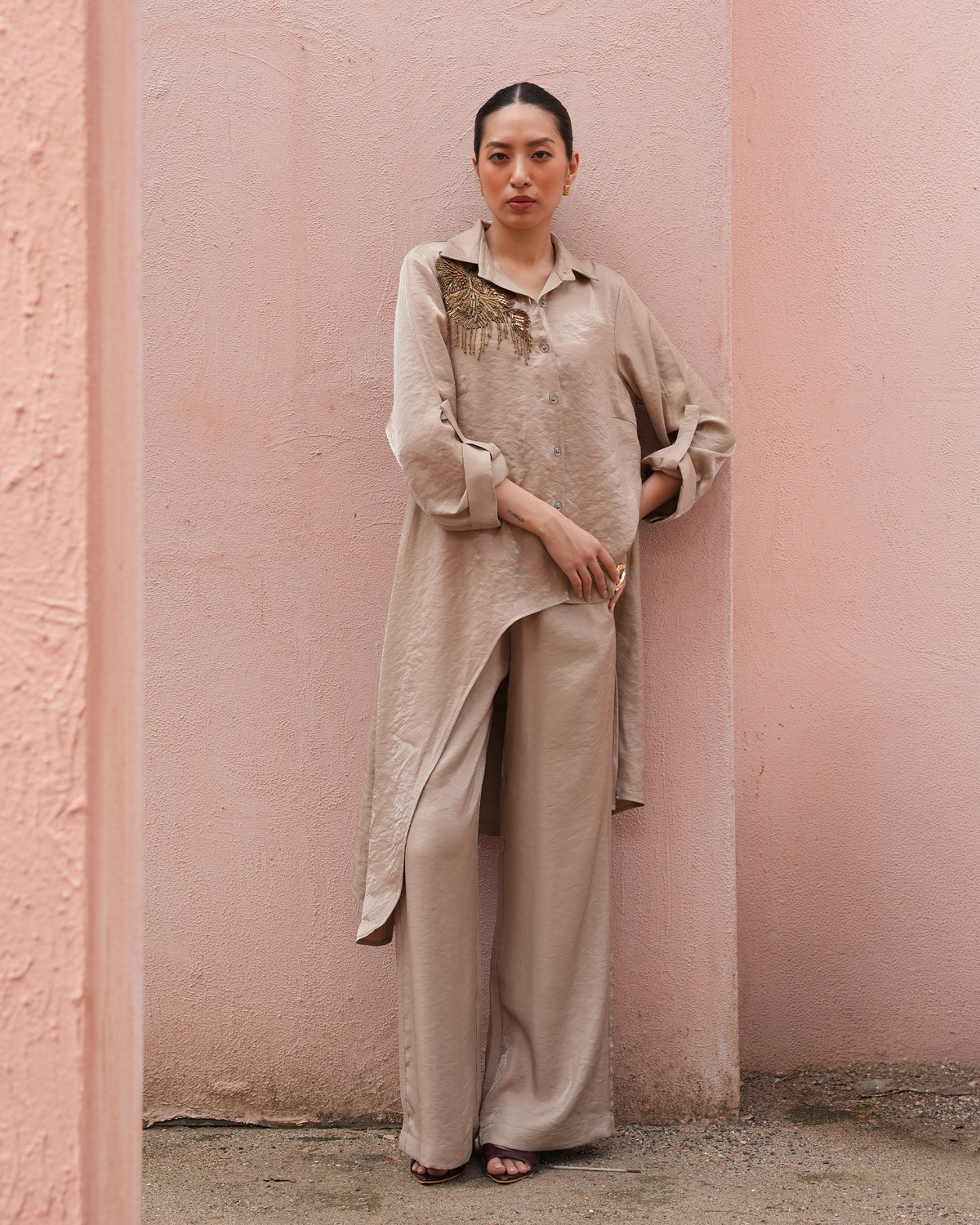 A woman stands against a textured pink wall wearing a matching beige lounge set consisting of a long-sleeve button-up shirt with embroidered detailing on the chest and wide-leg trousers.