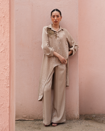 A woman stands against a textured pink wall wearing a matching beige lounge set consisting of a long-sleeve button-up shirt with embroidered detailing on the chest and wide-leg trousers.