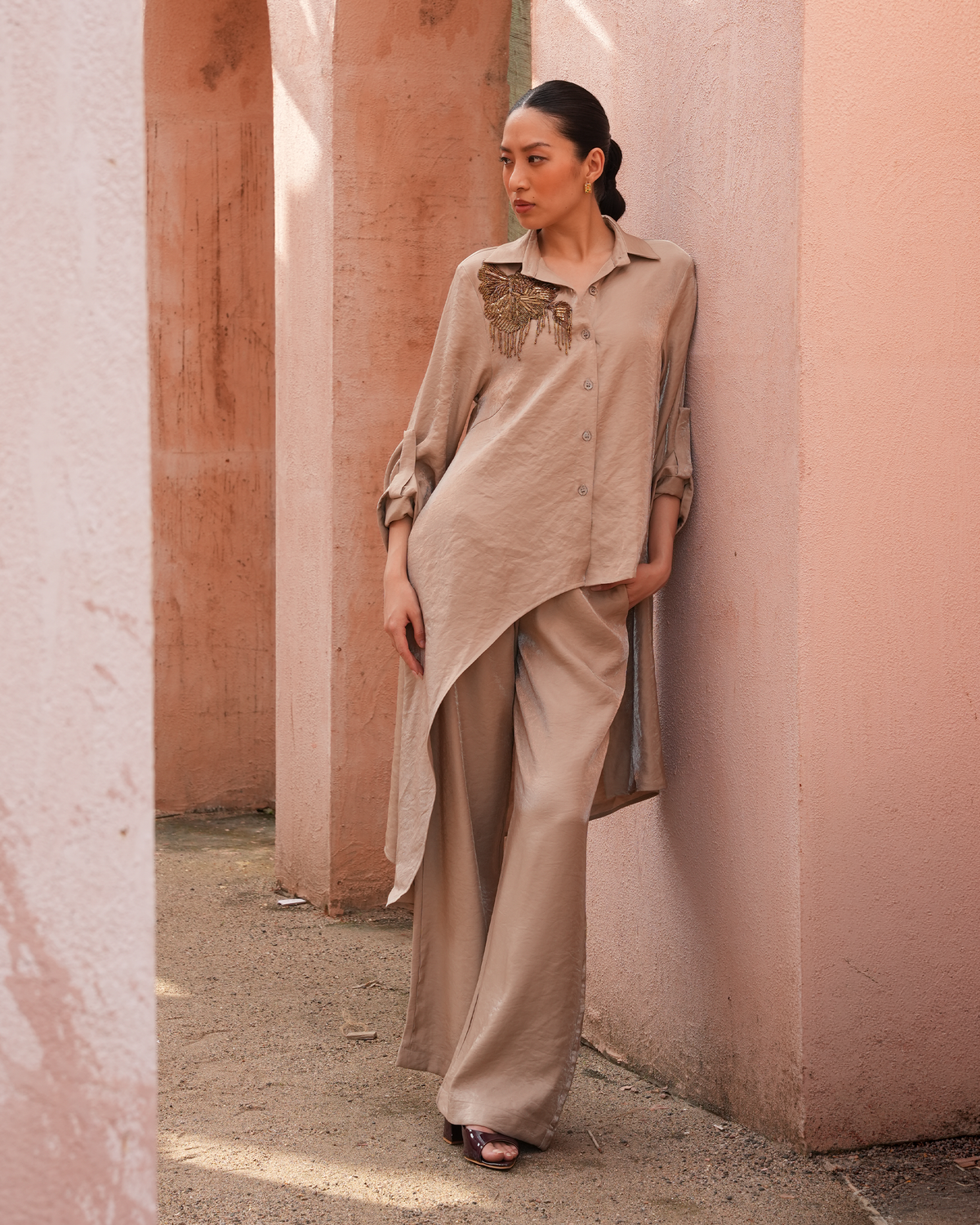 A woman stands in a narrow outdoor walkway between pink stucco columns, wearing a loose beige long-sleeve shirt with a decorative embroidered yoke and matching wide-leg trousers.