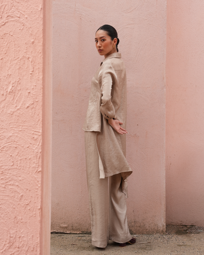 A woman stands in a narrow pink-walled corridor wearing a light beige matching outfit with a long-sleeve jacket and wide-leg pants.