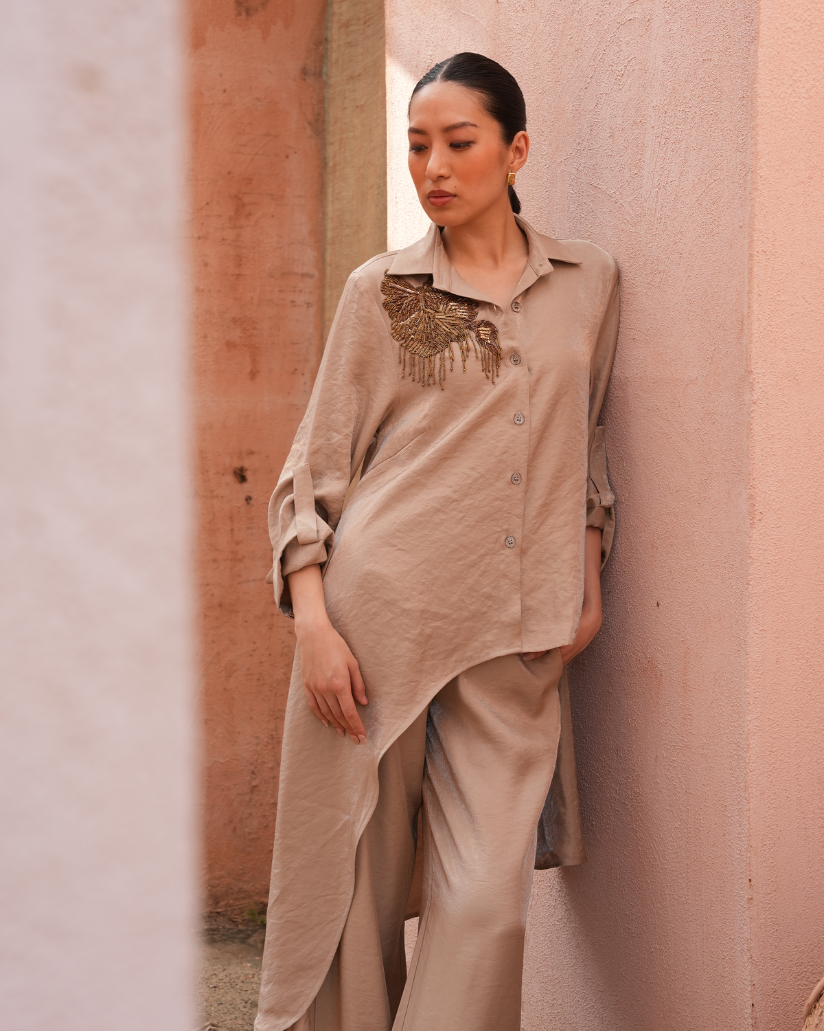 A woman leans against a textured pink wall wearing a beige linen button-up shirt with brown embroidered detailing on the chest and matching beige wide-leg trousers.