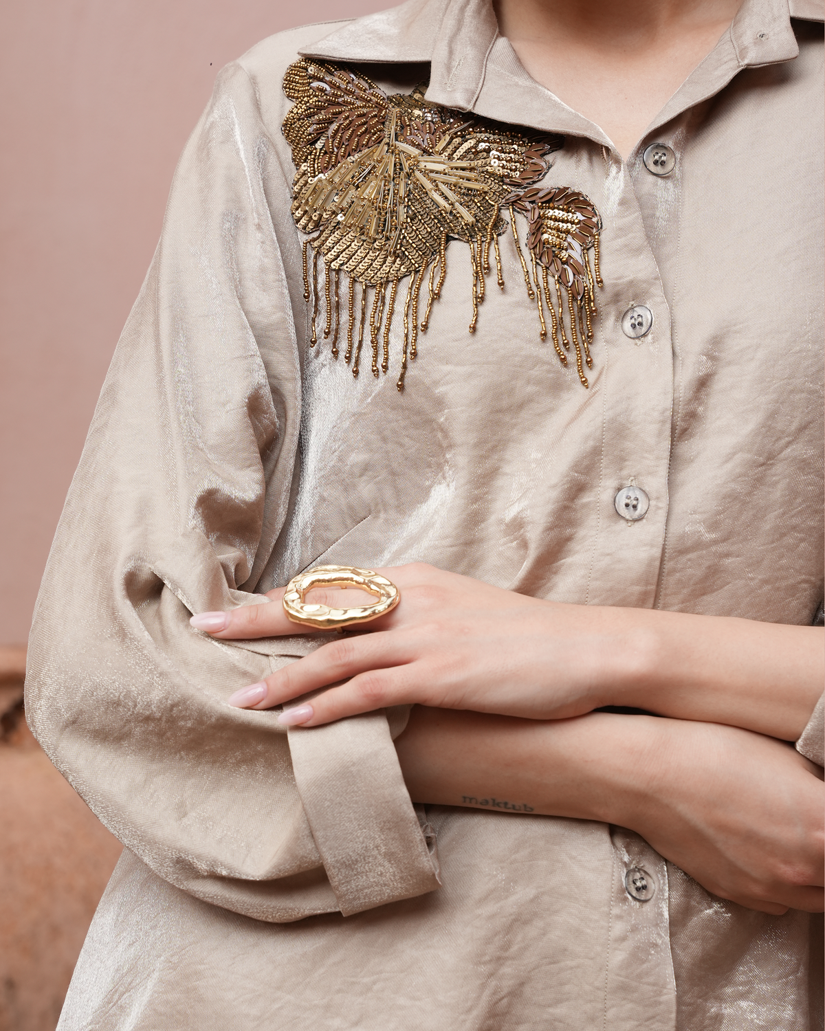 A close-up of a person wearing a beige linen button-up shirt with a large gold beaded butterfly embellishment on the chest. The person is also wearing a chunky gold ring on one hand, with the arms crossed.