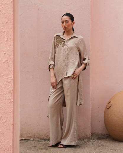 A woman stands barefoot against a textured pink wall, wearing a loose beige satin-like shirt and matching wide-leg pants with gold embroidery on the chest.