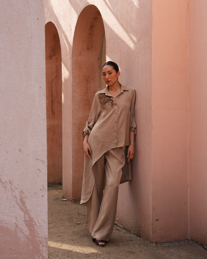 A woman stands in a dusty pink architectural setting with arches, wearing a loose taupe-beige button-up shirt with a textured chest detail and matching wide-leg trousers.