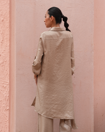 A woman shown from the back wearing a light beige linen long coat over matching trousers, standing against a textured pink wall.